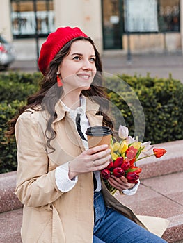 Beautiful young woman with coffee to go and spring tulips flowers bouquet at city street. Happy girl walking outdoors. Spring