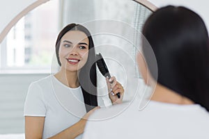 Beautiful young woman combing her hair in the morning, looking in a big round mirror