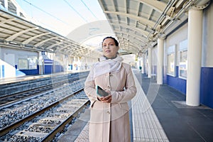 Beautiful young woman commuter holding mobile phone, waiting to board the train, on the platform of railway station