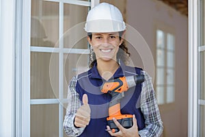 Beautiful young woman contractor with drill showing thumbs up