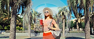 Beautiful young woman with cup of coffee in summer park wearing backpack on palm tree background