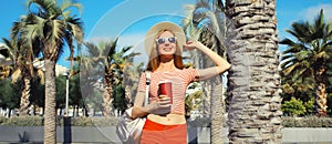 Beautiful young woman with cup of coffee in summer park wearing backpack on palm tree background