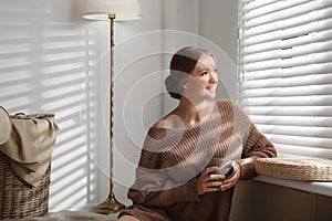 Beautiful young woman with cup of hot drink near window at home