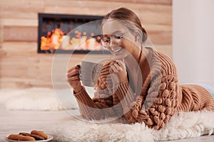 Beautiful young woman with cup of hot drink on rug. Winter atmosphere