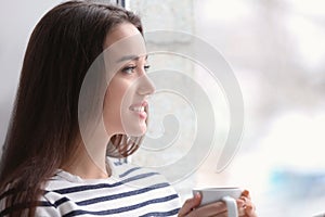 Beautiful young woman with cup of hot tea near window at home