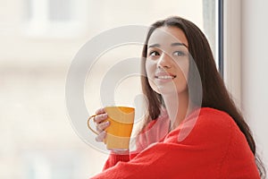 Beautiful young woman with cup of hot tea near window at home