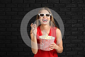 Beautiful young woman with cup of popcorn and 3D cinema glasses against dark brick wall