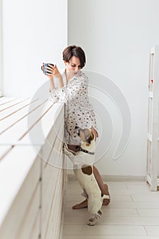 Calm and coziness. Beautiful young woman with cup of tea and dog standing near the window at home