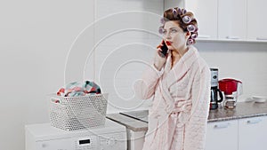 A beautiful young woman with curlers on her head stands next to the washing machine. She`s talking on her smartphone