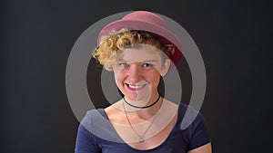 Beautiful young woman with curly blonde hair in hat smiling at camera, standing isolated on black background