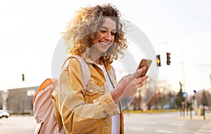 Beautiful young woman with curly hair smiling and using smartphone