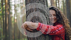 Beautiful young woman with curly hair wearing bright shirt is hugging tree enjoying nature and smiling with closed eyes