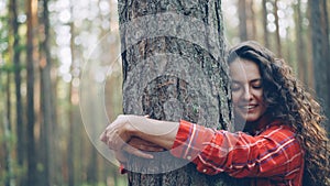 Beautiful young woman with curly hair wearing bright shirt is hugging tree enjoying nature and smiling with closed eyes