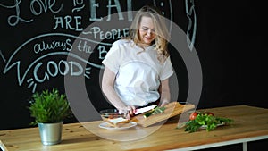 Beautiful young woman cuts green cucumber with kitchen knife on round pieces on wooden cutting board and prepares