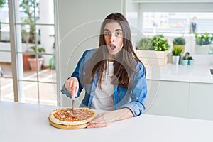 Beautiful young woman cutting a tasty pizza slice using a cutter scared in shock with a surprise face, afraid and excited with