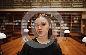 Beautiful young woman in dark clothes stands in a public library and poses for the camera on a background of shelves with books