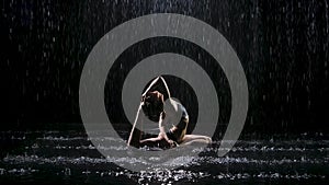 Beautiful young woman with dark hair in dove pose under the streams of rain. The studio light reflects on the water and