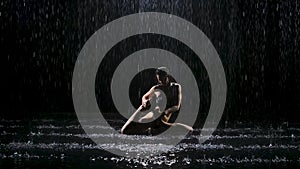 Beautiful young woman with dark hair performs complex asanas under the streams of rain. The studio light reflects on the