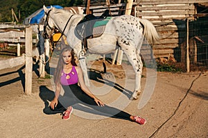 Beautiful young woman with dark hair in sportive clothes posing with white horse in stable