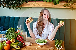 Beautiful young woman decides eating hamburger or apple in kitchen. Cheap junk food vs healthy diet