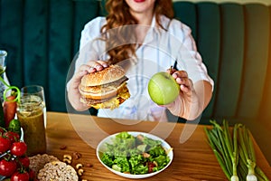 Beautiful young woman decides eating hamburger or apple in kitchen. Cheap junk food vs healthy diet