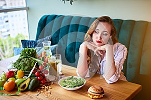 Beautiful young woman decides eating hamburger or vegetables  in kitchen. Cheap junk food vs healthy diet