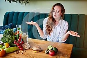 Beautiful young woman decides eating hamburger or vegetables  in kitchen. Cheap junk food vs healthy diet