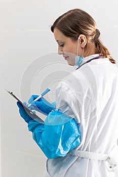 Beautiful young woman doctor in a medical gown, mask and blue gloves with a stethoscope writes down a medical history