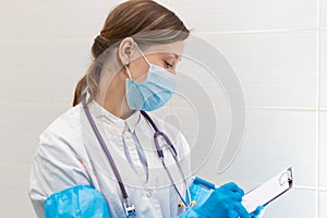 Beautiful young woman doctor in a medical gown, mask and blue gloves with a stethoscope writes down a medical history in a hospita