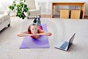 Beautiful young woman doing exercise for back on floor at home, online training on laptop computer, copy space.
