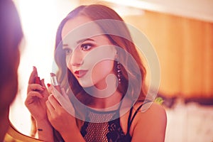 A beautiful young woman is doing herself a makeover. Girl with cerly hair doing evening makeup using lipstick in front of mirror i