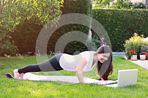 Beautiful young woman doing sport in her garden outdoors following guide of online tutorial or trainer on laptop. Healthy lifestyl