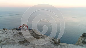 Beautiful young woman is doing yoga stretcing on the top of mountain with sea view at sunset. Aerial footage.