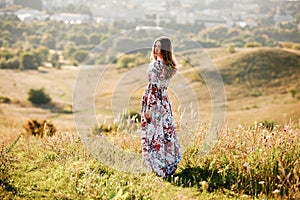 Beautiful young woman in dress in floral print walking in the field at sunset. stylish romantic girl with long hair have a good