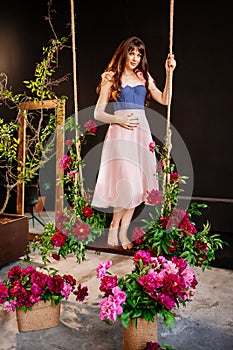 a beautiful young woman in a dress stands on a swing in peony flowers.