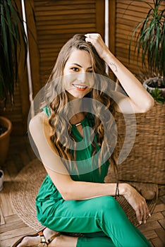 A beautiful young woman dressed in a green jumpsuit holds her hand on her head and smiles