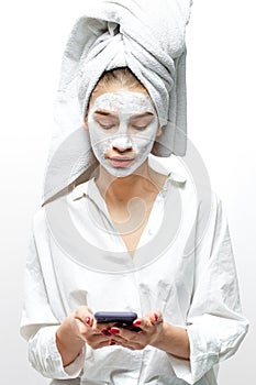 Beautiful young woman dressed in white clothes with a white towel on her hair and cosmetic mask on her face holds mobile