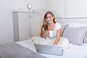 Beautiful young woman drinking coffee at home in her bed wearing a cozy t shirt while working on her laptop. Checking email in the