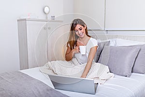 Beautiful young woman drinking coffee at home in her bed wearing a cozy t shirt while working on her laptop. Checking email in the