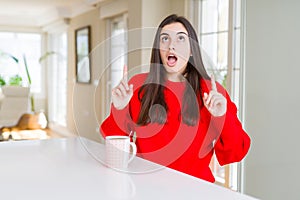 Beautiful young woman drinking a cup of black coffee amazed and surprised looking up and pointing with fingers and raised arms