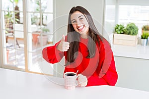 Beautiful young woman drinking a cup of black coffee doing happy thumbs up gesture with hand