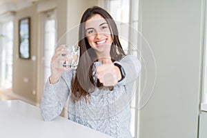 Beautiful young woman drinking a fresh glass of water happy with big smile doing ok sign, thumb up with fingers, excellent sign