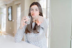 Beautiful young woman drinking a fresh glass of water pointing with finger to the camera and to you, hand sign, positive and