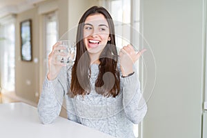 Beautiful young woman drinking a fresh glass of water pointing and showing with thumb up to the side with happy face smiling