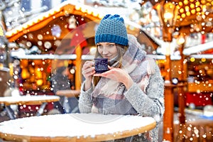 Beautiful young woman drinking hot punch, mulled wine on Christmas market. Happy girl in winter clothes with lights on