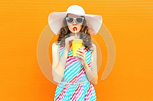 Beautiful young woman drinking juice in summer straw hat, colorful striped dress on orange wall