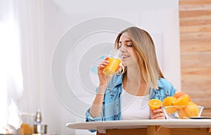 Beautiful young woman drinking orange juice at table indoors, space for text