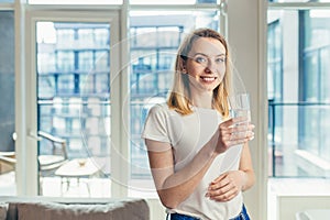 Happy beautiful young woman drinking water. Smiling caucasian female blond model holding transparent glass in her hand.