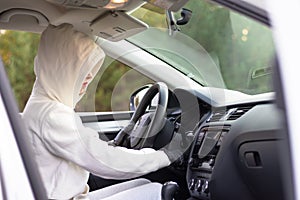 A beautiful young woman driver in a white jacket with a hood and black gloves sits at the wheel of a car. Selective focus. Portrai