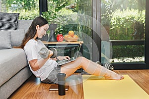 Beautiful young woman with dumbbell watching fitness video tutorials online on laptop while sit down on floor in living room